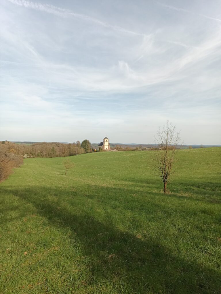 Landschaft mit Kapelle in der Ferne, umgeben von grünen Wiesen und Hügeln