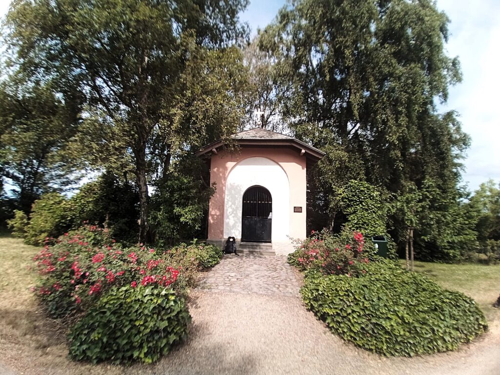 Außenansicht der Broichhofkapelle in Wachtberg, umgeben von Bäumen und Rosensträuchern.