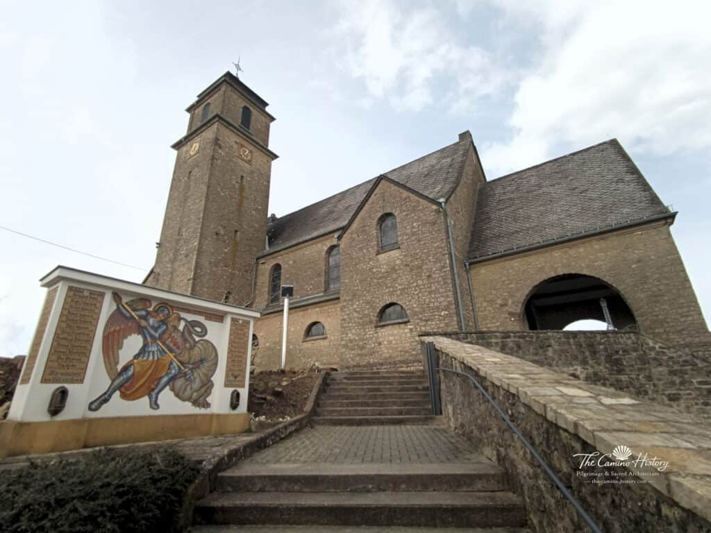 Ansicht der Pfarrkirche St. Johannes der Täufer in Borg mit Kirchturm und einem Mosaik des Erzengels Michael