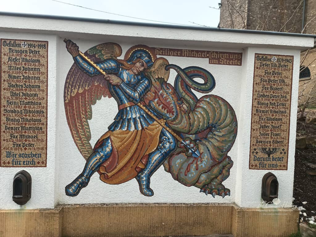 Mosaik des Erzengels Michael, der einen Drachen bekämpft, eingerahmt von den Namen der Gefallenen der Weltkriege an einer Gedenkwand neben der Pfarrkirche St. Johannes der Täufer in Borg.