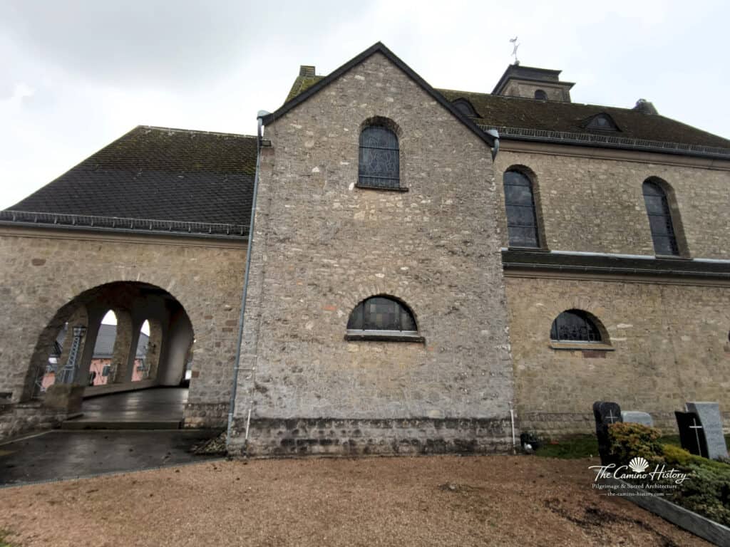 Seitliche Ansicht der Pfarrkirche St. Johannes der Täufer in Borg, mit steinernen Fassaden, Rundbogenfenstern und einem überdachten Durchgang im Vordergrund.