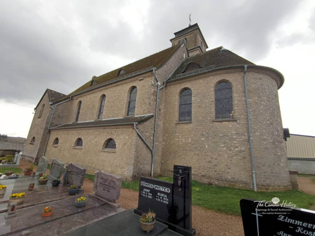 Rückansicht der Pfarrkirche St. Johannes der Täufer in Borg mit abgerundeter Apsis, steinernen Wänden und Grabsteinen im Vordergrund.