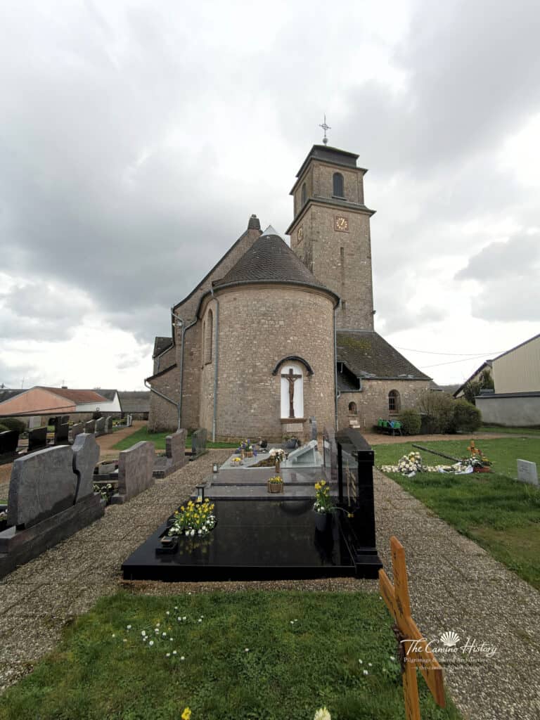 Rückansicht der Pfarrkirche St. Johannes der Täufer in Borg, umgeben von einem Friedhof mit blühenden Gräbern und einem Kruzifix.
