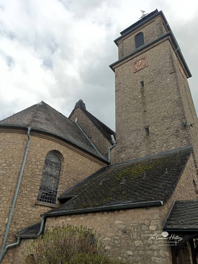 Nahaufnahme des Kirchturms der Pfarrkirche St. Johannes der Täufer in Borg, umgeben von einem steinernen Anbau und mit einem markanten Wetterhahn auf der Turmspitze.