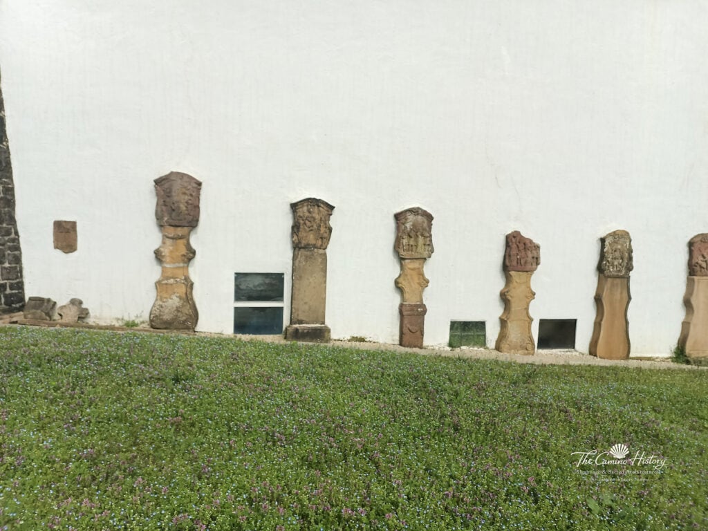 Reihe von historischen Grabkreuzen aus Stein, aufgestellt entlang der weiß verputzten Außenwand der Pfarrkirche St. Johannes der Täufer in Borg.