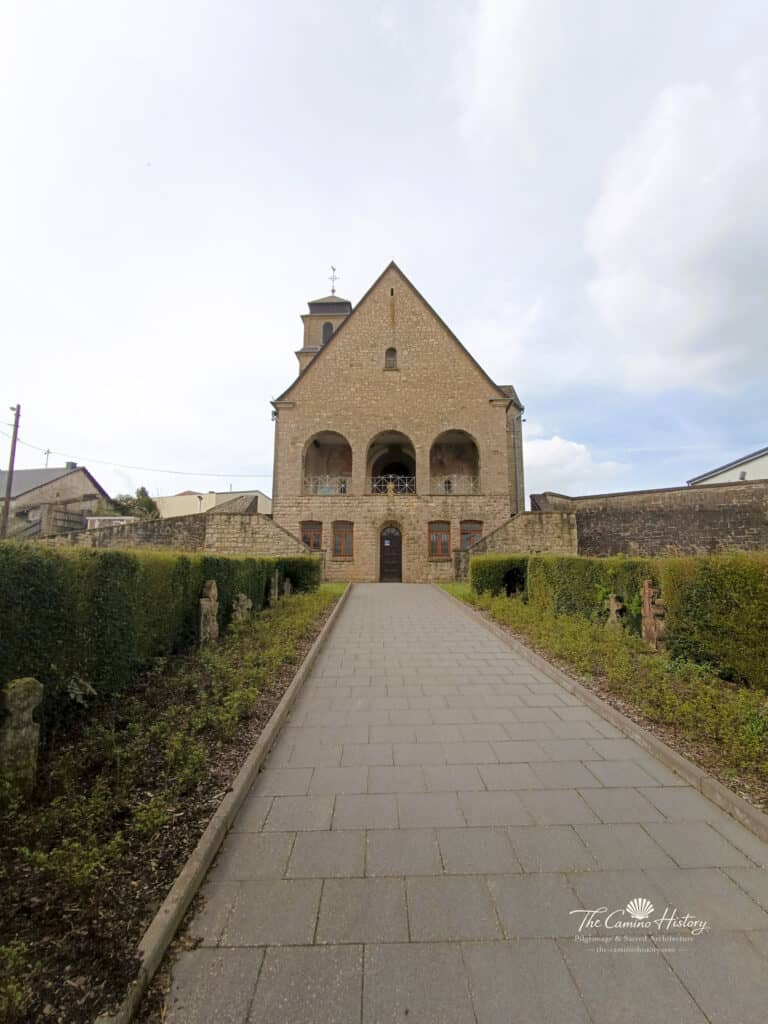Hauptweg und Eingangsbereich der Pfarrkirche St. Johannes der Täufer in Borg, umgeben von gepflegten Hecken und historischen Grabsteinen.