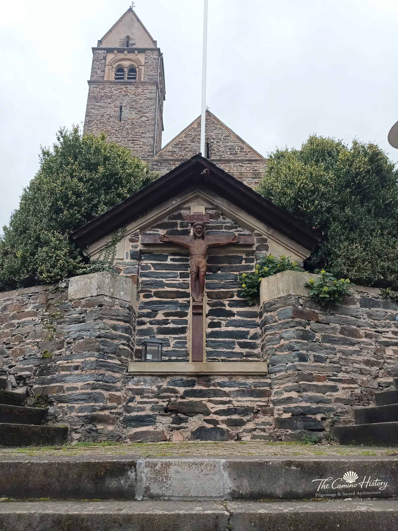 Kruzifix an der Treppe zu St. Peter und Paul in Traben; dahinter die Schieferfassade mit Kirchturm.