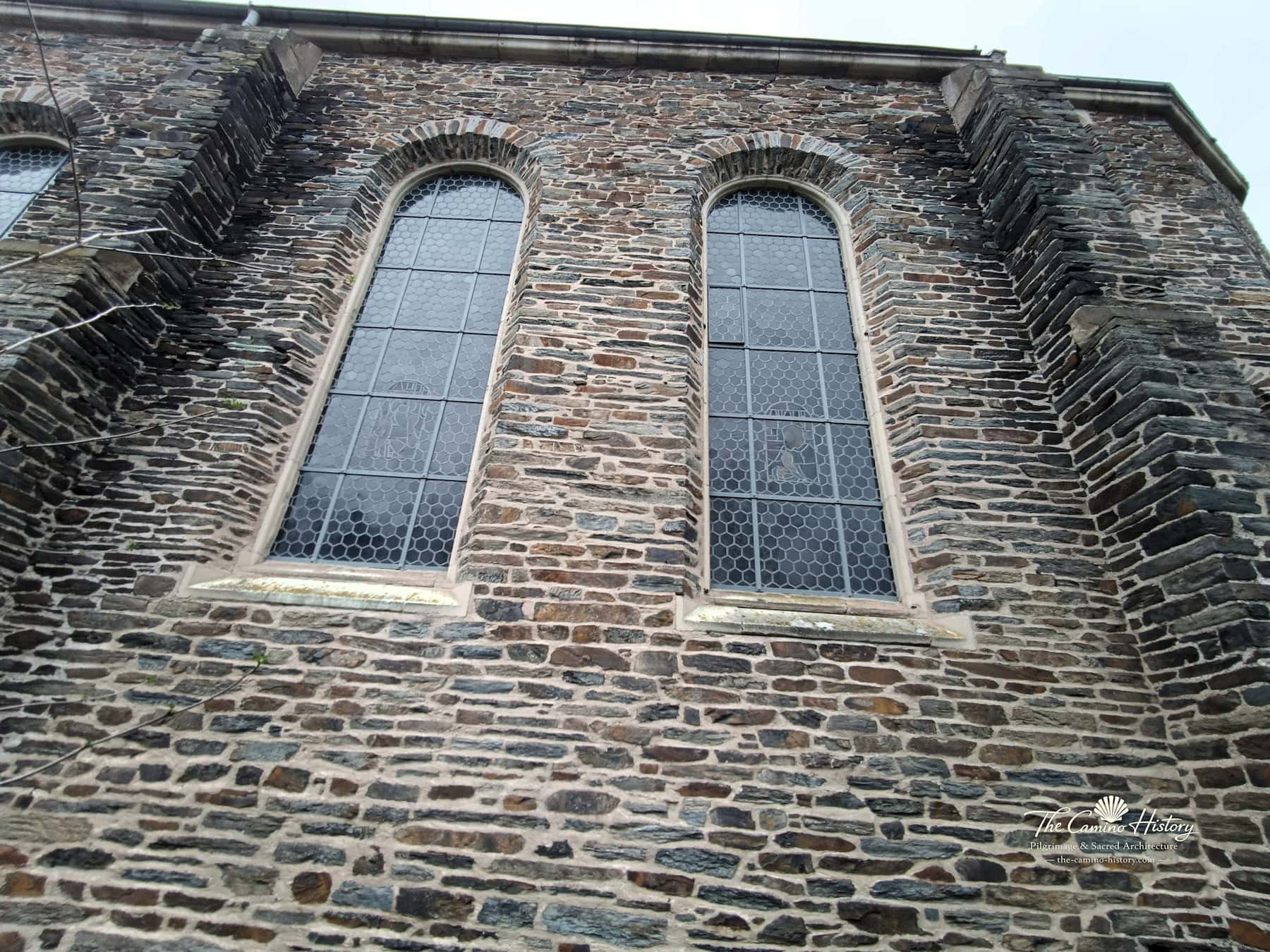 Rundbogige Glasfenster der Kirche St. Peter und Paul in Traben zwischen markanten Strebepfeilern im Schiefermauerwerk.