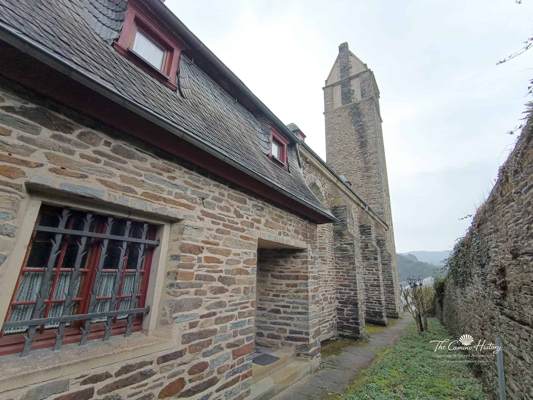 Seitlicher Weg entlang der Kirche St. Peter und Paul in Traben mit Schieferdach, Strebepfeilern und hohem Turm.